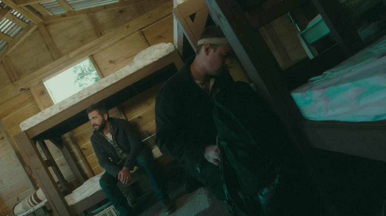 dutch angle shots example from The Sacrament: A wooden cabin interior features exposed beams and built-in wooden bunk beds. One man sits on a bed whil