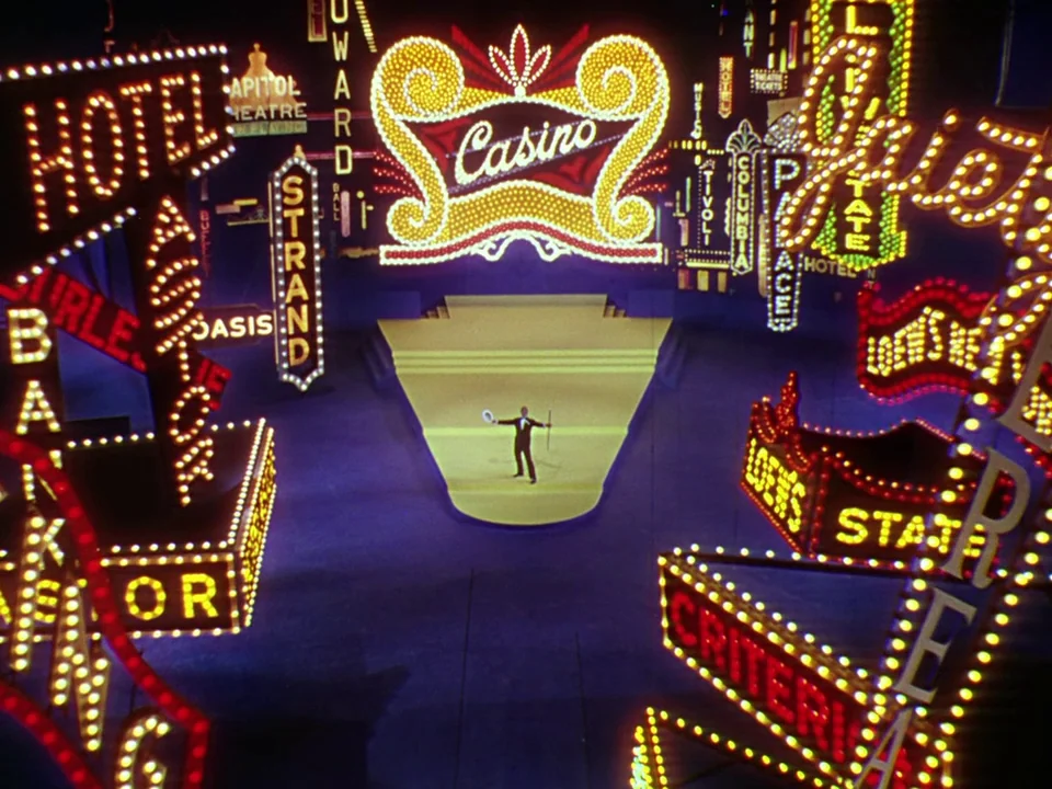 Singin’ in the Rain (1952) movie still: wide — A dark theatrical stage features an elevated platform surrounded by towering vintage neon signage. A…