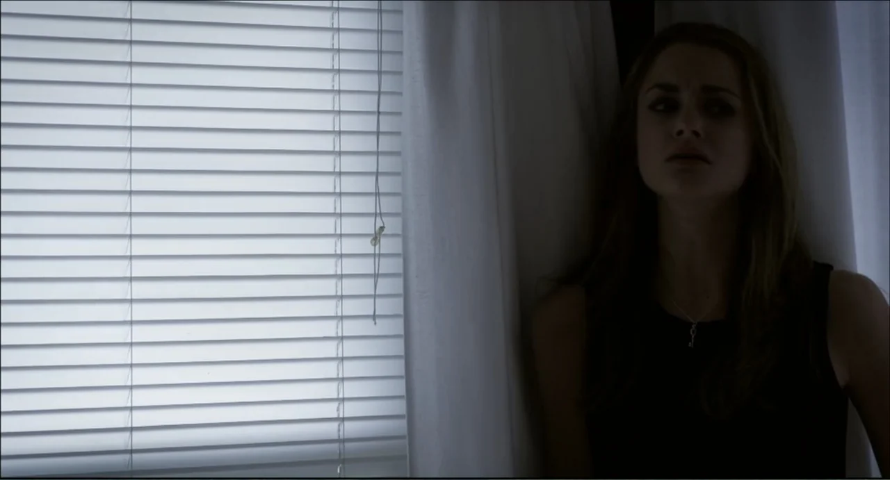 The Secret Village (2013) movie still: medium — An interior space with a window covered by closed horizontal blinds at night. A young woman stands…