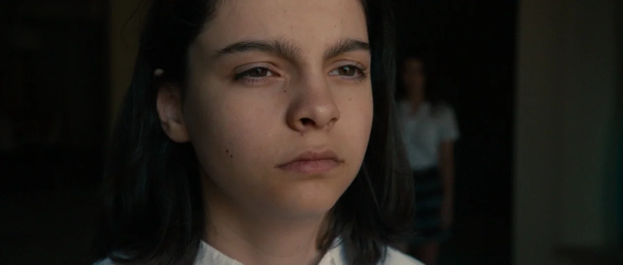 The Purge (2013) movie still: closeup — A dark indoor setting provides a backdrop for a young girl in white clothing. She stands in the foreground…