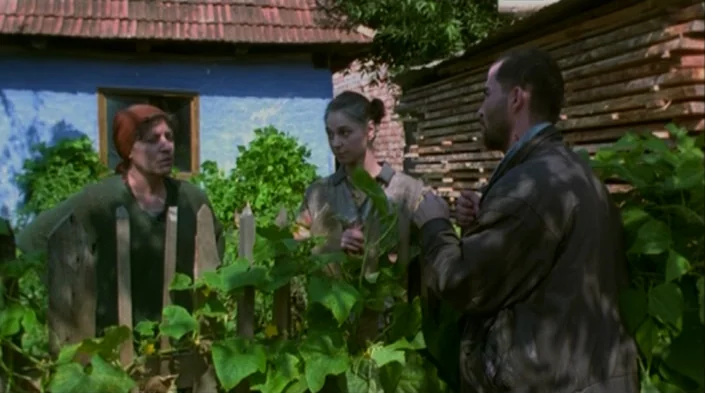 Code Unknown (2000) movie still: medium — A rural backyard features a blue-walled house and a stacked pile of lumber. Three adults stand around a…