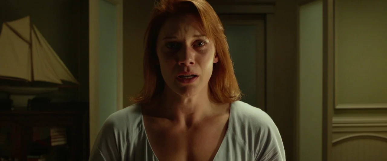 Oculus (2013) movie still: closeup — A dimly lit room contains a model sailboat on a dark wooden shelf. A woman with red hair stares directly at the…