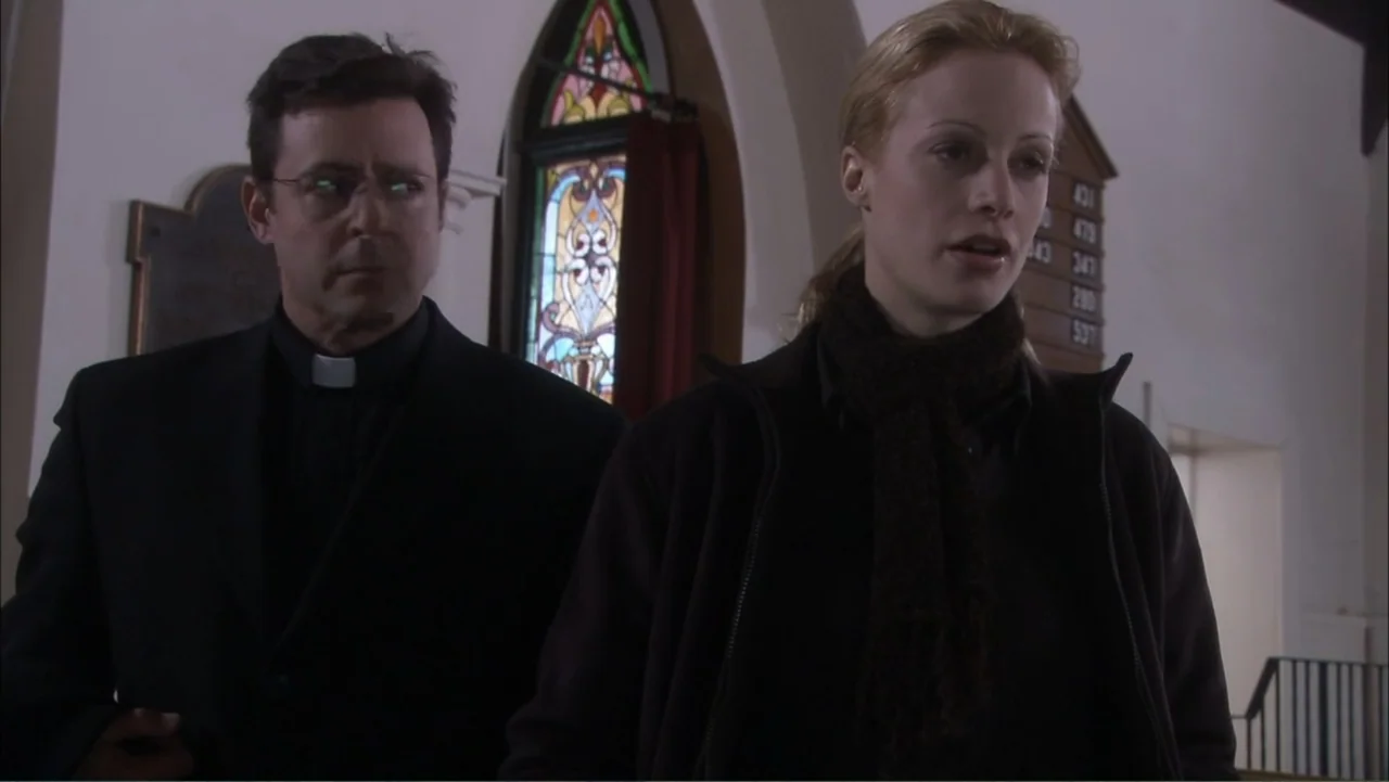 O código oculto (2025) movie still: medium — The interior of a church features a wooden hymn board and stained glass window. A priest stands next to…