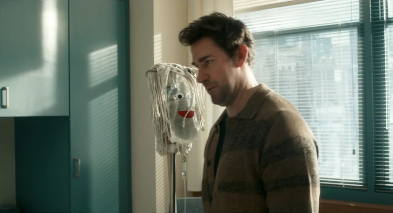 IF (2024) movie still: medium — A hospital room with teal cabinets and blinds covering the window. A man stands beside an IV pole with a decorated…