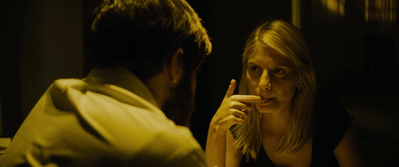 Enemy (2013) movie still: medium — A dimly lit room holds a man in a white shirt and a blonde woman. The woman sits opposite the man, holding a…