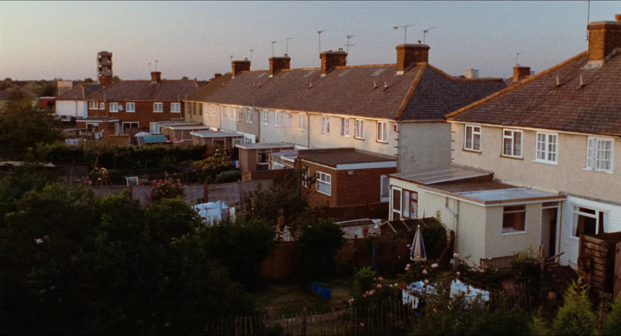 Life Is Sweet (1990) movie still: establishing — A row of terraced houses with tiled roofs stretches across a quiet suburban street. Lush green…