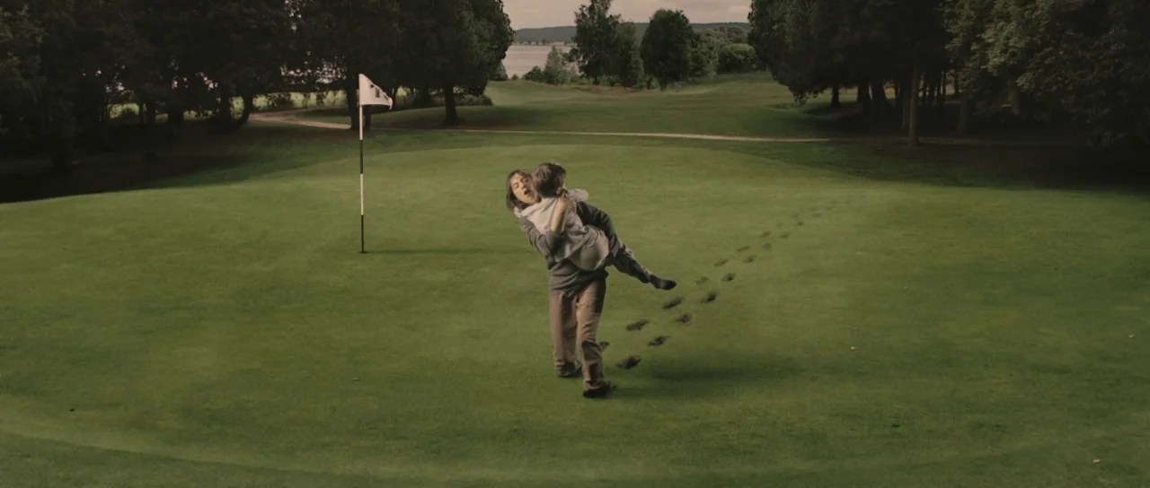 Melancholia (2011) movie still: wide — A wide, lush green golf course features a flag pin near a large grass area. A woman carries a child in her…