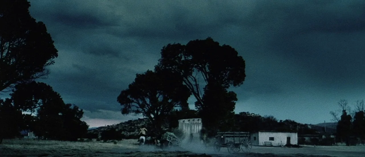 Renegade (2004) movie still: wide — An arid, dusty settlement sits under a dark, thick layer of storm clouds. A small wooden wagon travels along a…