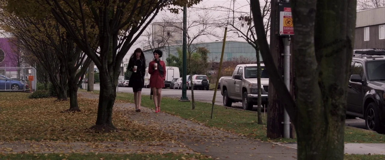 American Mary (2012) movie still: fullbody — A suburban sidewalk is covered in fallen autumn leaves with industrial buildings in the background. Two…