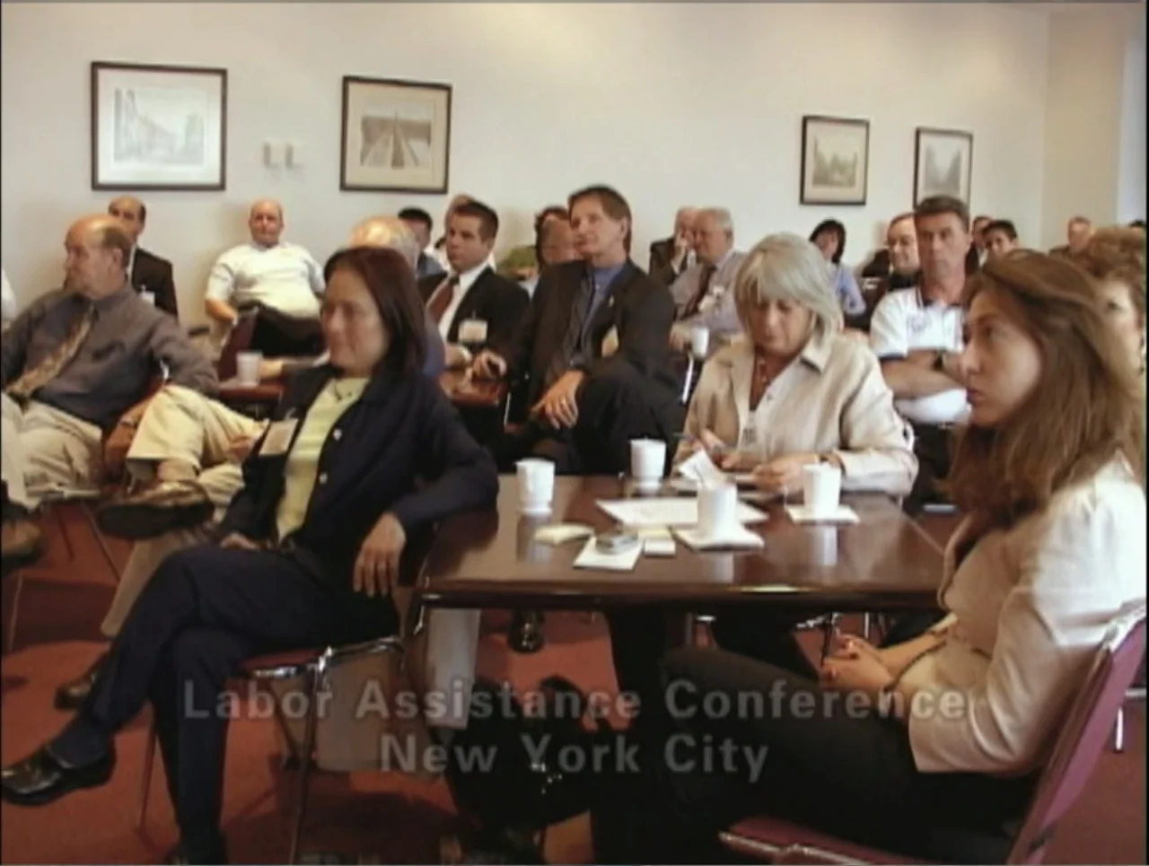 Addiction (2024) movie still: medium — A beige-walled conference room features several wooden tables and framed pictures on the wall. A group of…