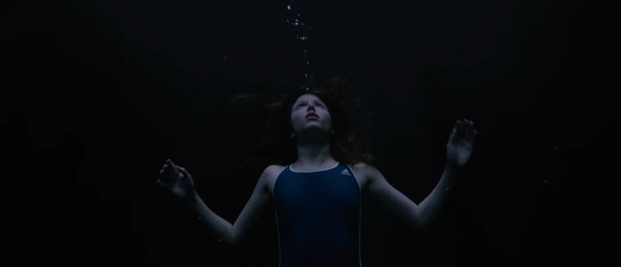 Thelma (2017) movie still: medium — A person is submerged in deep dark water looking upward toward the surface. Bubbles rise from behind her head as…