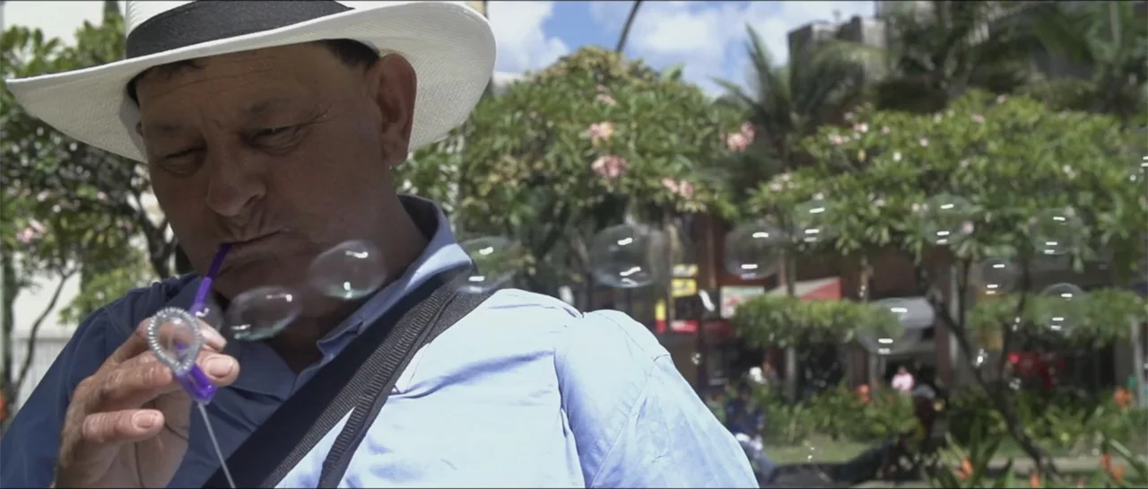 10 Years with Bebe (2016) movie still: medium — A sunny public park with lush green foliage serves as the backdrop to a man. An older man in a straw…