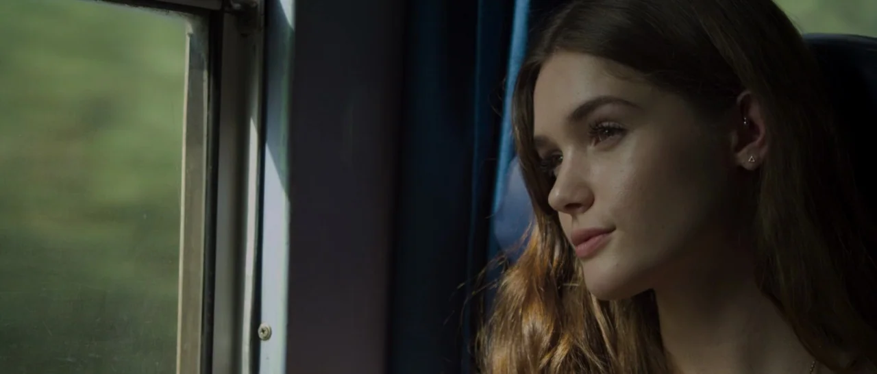 The Face of an Angel (2014) movie still: closeup — The interior of a moving passenger train with dark blue seat upholstery. A young woman sits by…