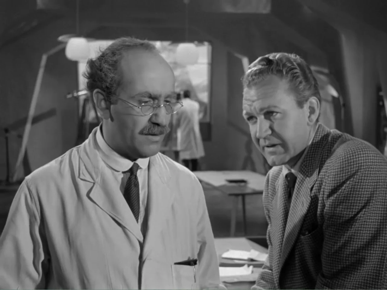 The Crawling Eye (1958) movie still: medium — A sterile laboratory space featuring hanging glass lamps and scattered papers on tables. A man in a…