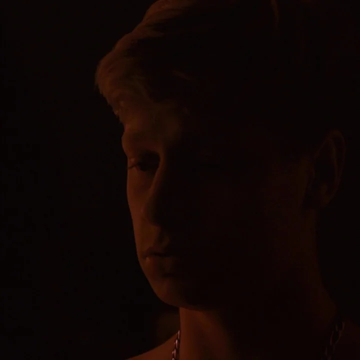 Mommy (2014) movie still: closeup — A dark space provides a minimal backdrop for a close view of a person. A young man stands with his head turned,…