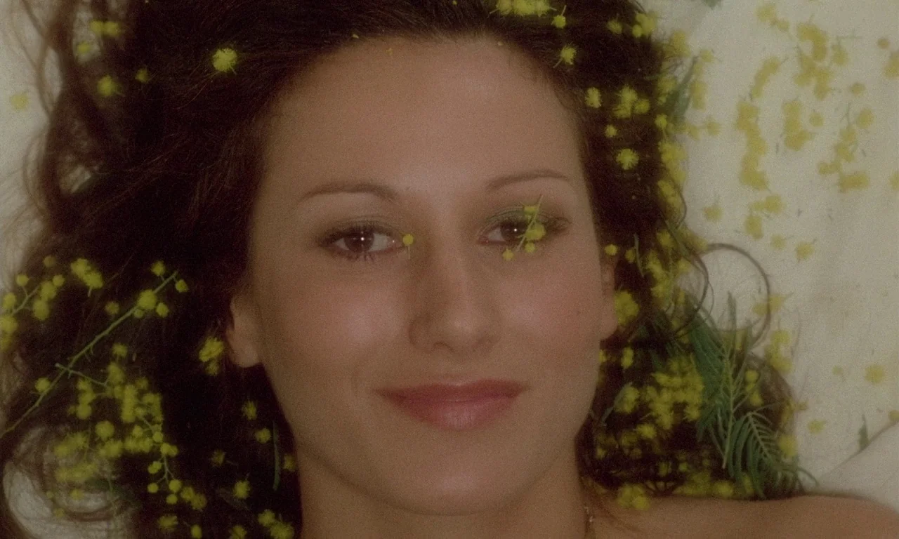 The Margin (1976) movie still: closeup — A woman lies on a white surface with yellow flowers scattered around her head. She faces the camera…