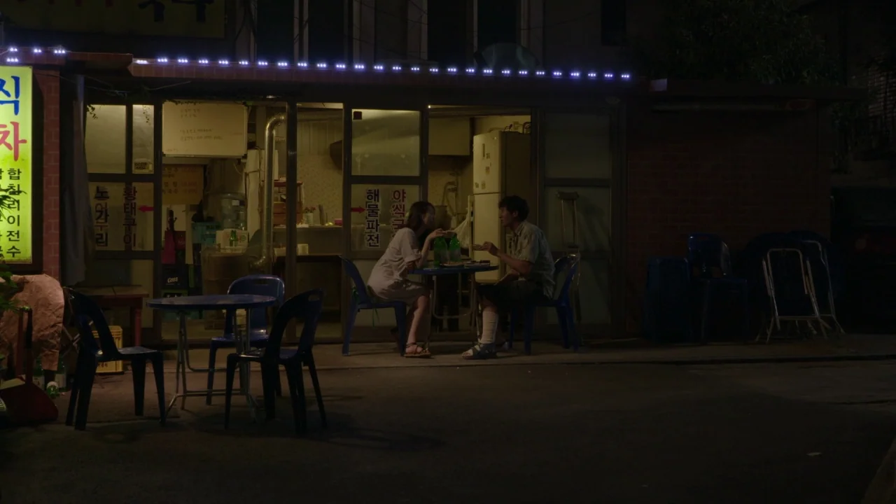 Yourself and Yours (2016) movie still: wide — A night view of a small Korean storefront with an outdoor seating area. A young man and woman sit at a…