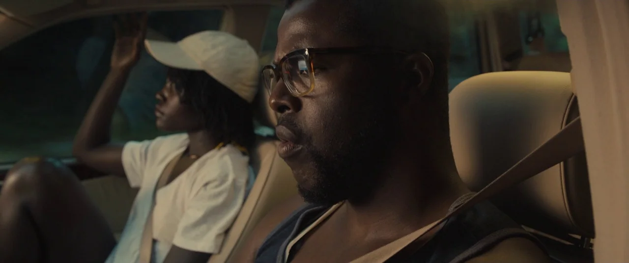 Us (2019) movie still: medium — The interior of a moving vehicle during daylight hours with tan upholstery visible. A man drives while a woman sits…