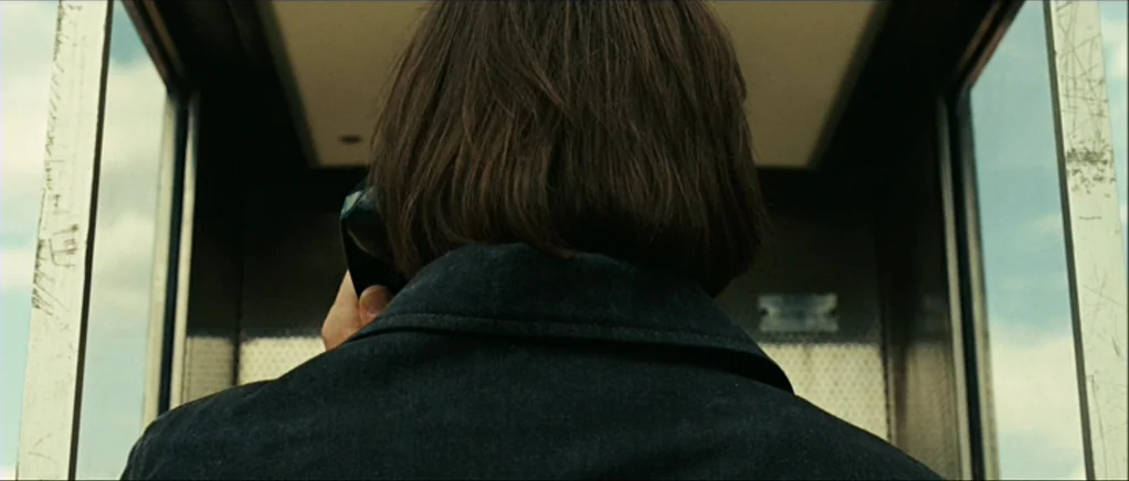 No Country For Old Men movie still: closeup — A metal-framed public enclosure features glass panels on the left and right sides. A man wearing a…