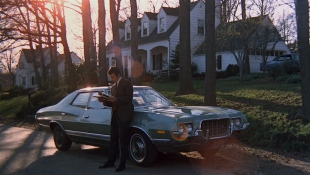 Sisters (1973) movie still: wide — A man stands next to a green sedan parked on a residential street. A two-story white house stands in the…