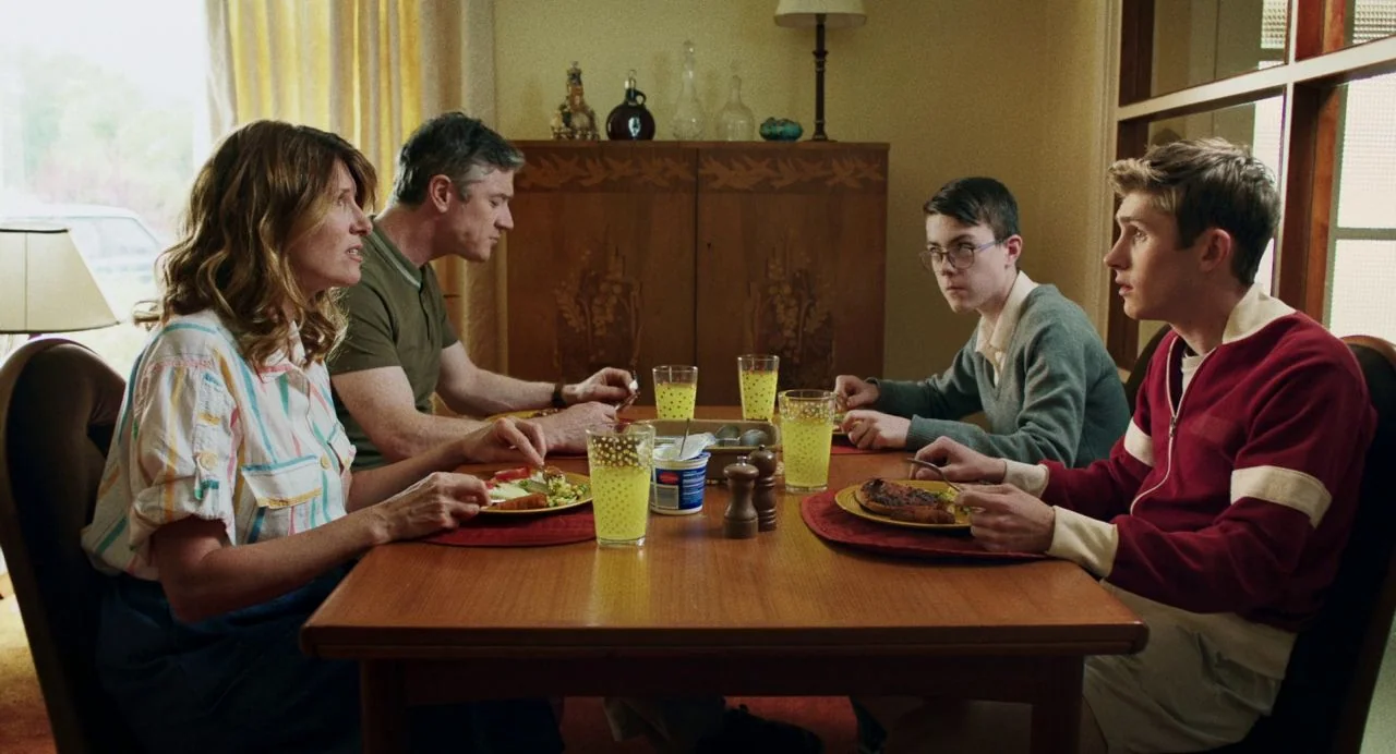 Dating Amber movie still: medium — A family sits around a wooden table in a modest suburban home with dated decor. Two parents and their two sons…