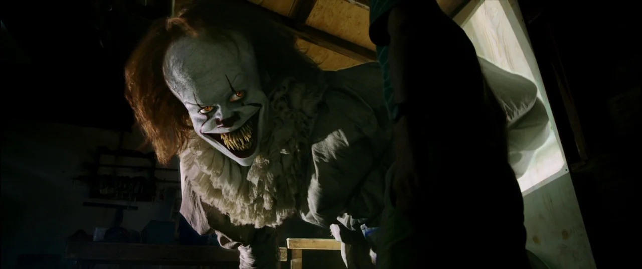 It (2017) movie still: medium — A dark wooden interior contains a clown leaning menacingly toward a partially visible boy. The clown displays sharp…