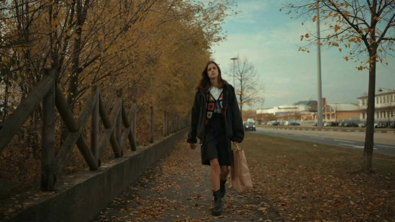 Amanda (2022) movie still: fullbody — A sidewalk runs parallel to a road bordered by autumn trees with yellow leaves. A young woman walks toward the…