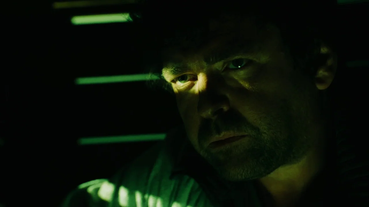 Saw III (2006) movie still: closeup — A man's face is partially illuminated in a dark, shadow-filled interior space. Horizontal light streaks from…