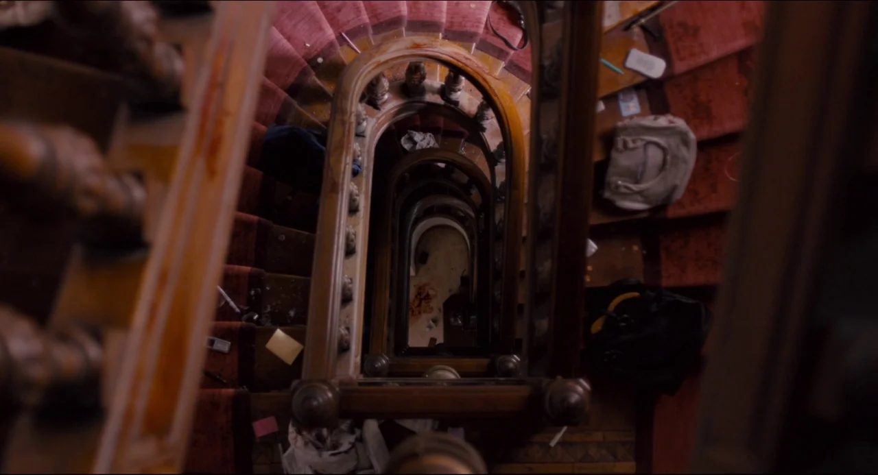 The Night Eats the World (2018) movie still: establishing — A top-down view looks down a spiraling wooden staircase with worn red carpet runners.…
