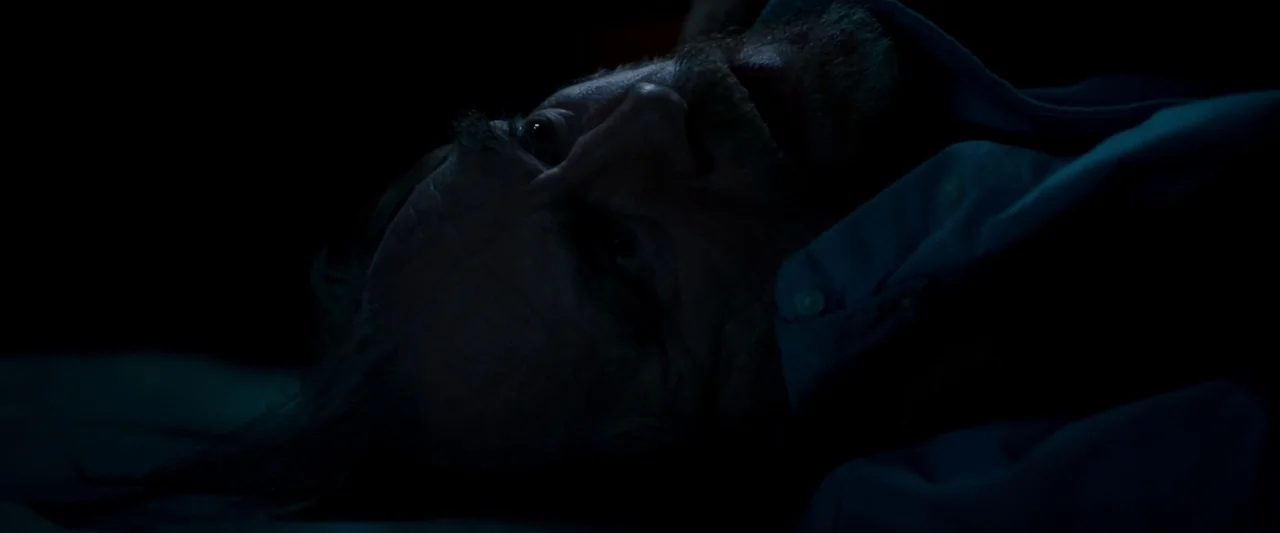 The Autopsy of Jane Doe (2016) movie still: closeup — A man lies on a white pillow in a dark room. He is positioned on his side and looks directly…