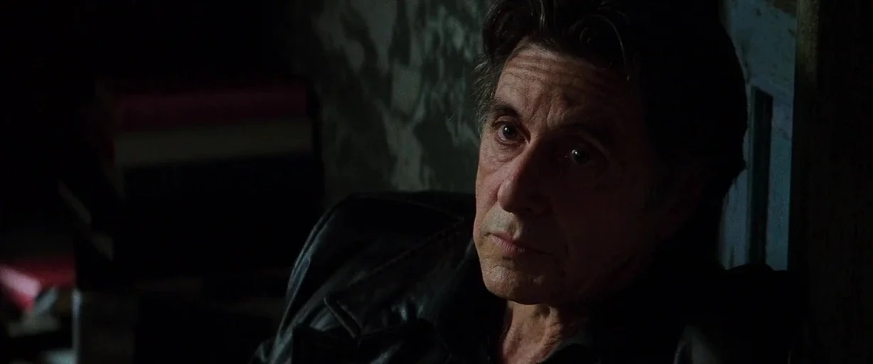 Insomnia (2002) movie still: closeup — A dimly lit room with textured dark walls behind an elderly man. The man sits motionless in a leather jacket…