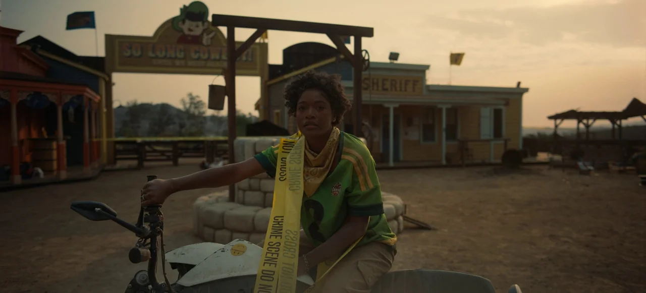 Nope (2022) movie still: medium — A dusty outdoor theme park set features a wooden sheriff building and gallows structure. A young woman sits on a…