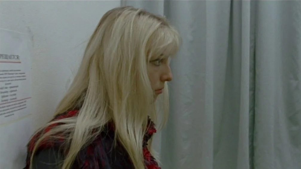 Import Export (2007) movie still: closeup — A confined area with white walls features a hanging grey privacy curtain. A young woman with blonde hair…
