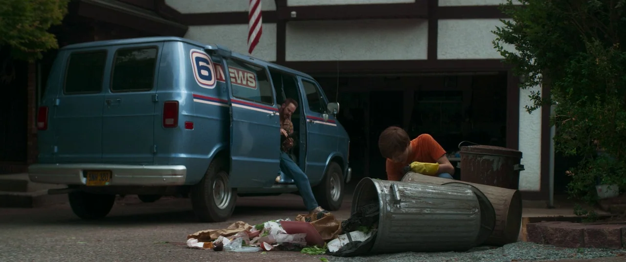 Summer of 84 (2018) movie still: wide — A suburban residential driveway features a light blue news van parked near a garage. A teenager in orange…