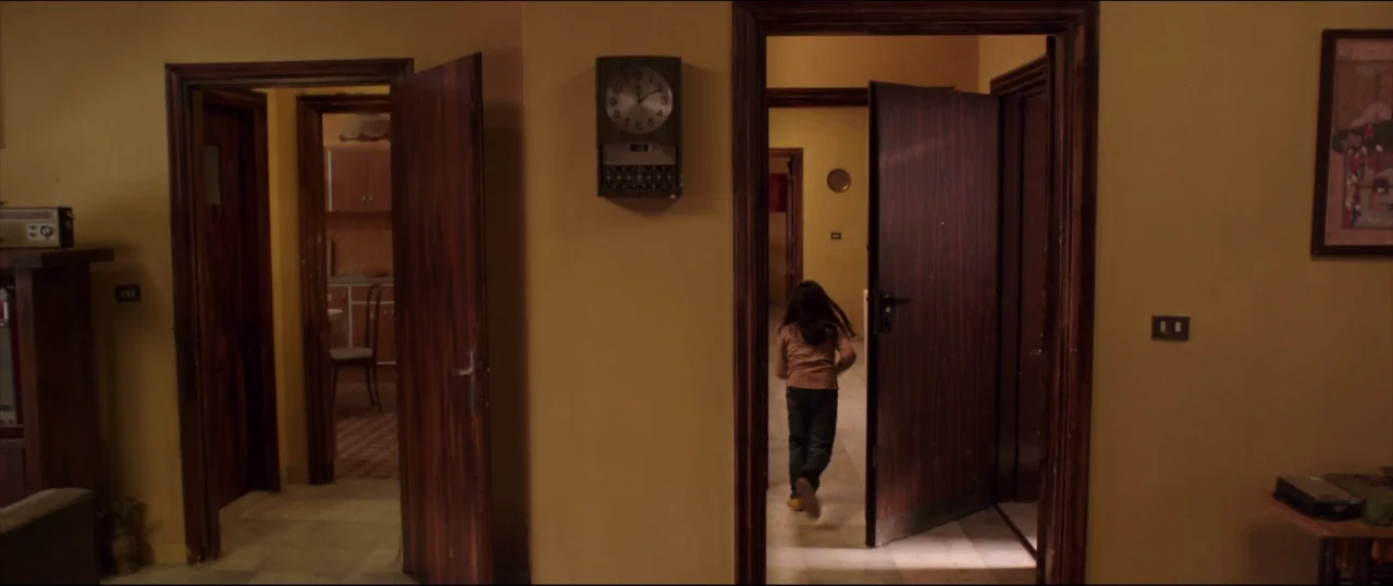 Under The Shadow (2016) movie still: wide — An interior space features yellow-painted walls, dark wooden doors, and a wall-mounted clock. A young…