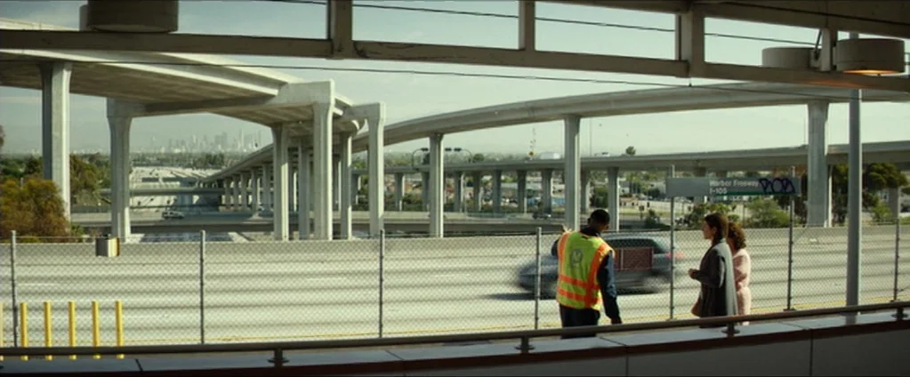 Cake (2014) movie still: wide — An elevated transit platform overlooks a sprawling concrete freeway interchange on a clear day. A transit worker…