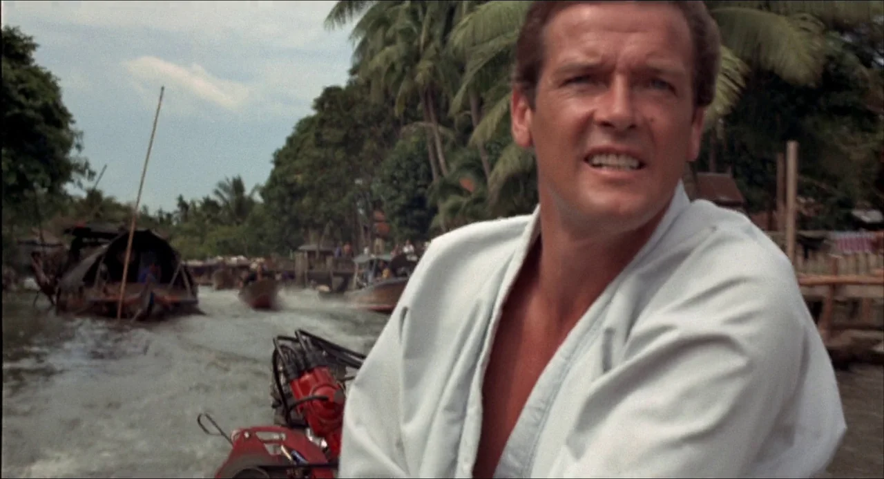 The Man With the Golden Gun (1974) movie still: medium — A man drives a motorized boat along a narrow waterway lined with palm trees. Several wooden…