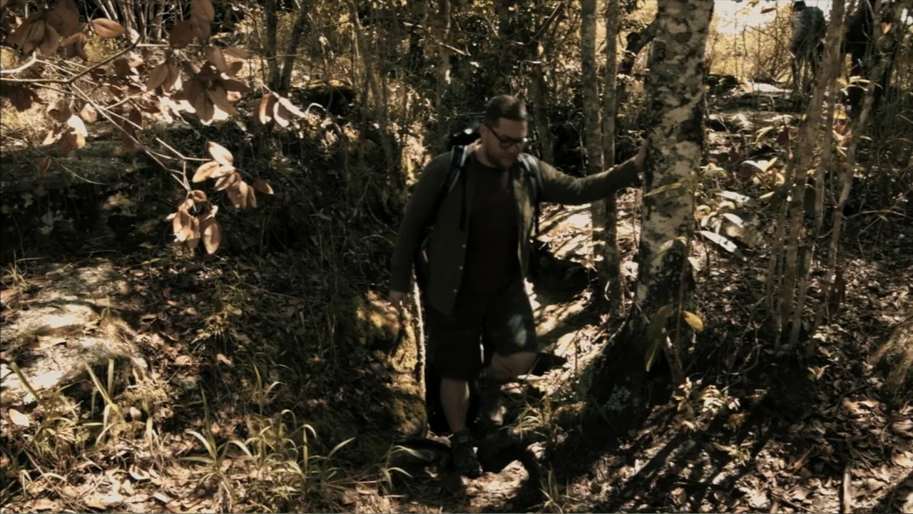 The Grotto (2022) movie still: fullbody — A man walks along a rocky path surrounded by dense trees and dried leaves. He moves forward while bracing…