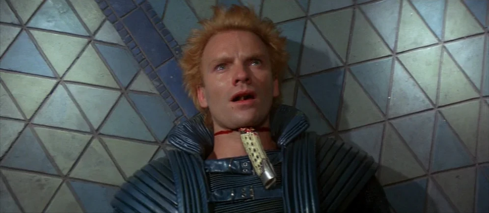 Dune (1984) movie still: closeup — A man lies against a wall covered in triangular blue and grey tiles. A metallic device is strapped tightly…