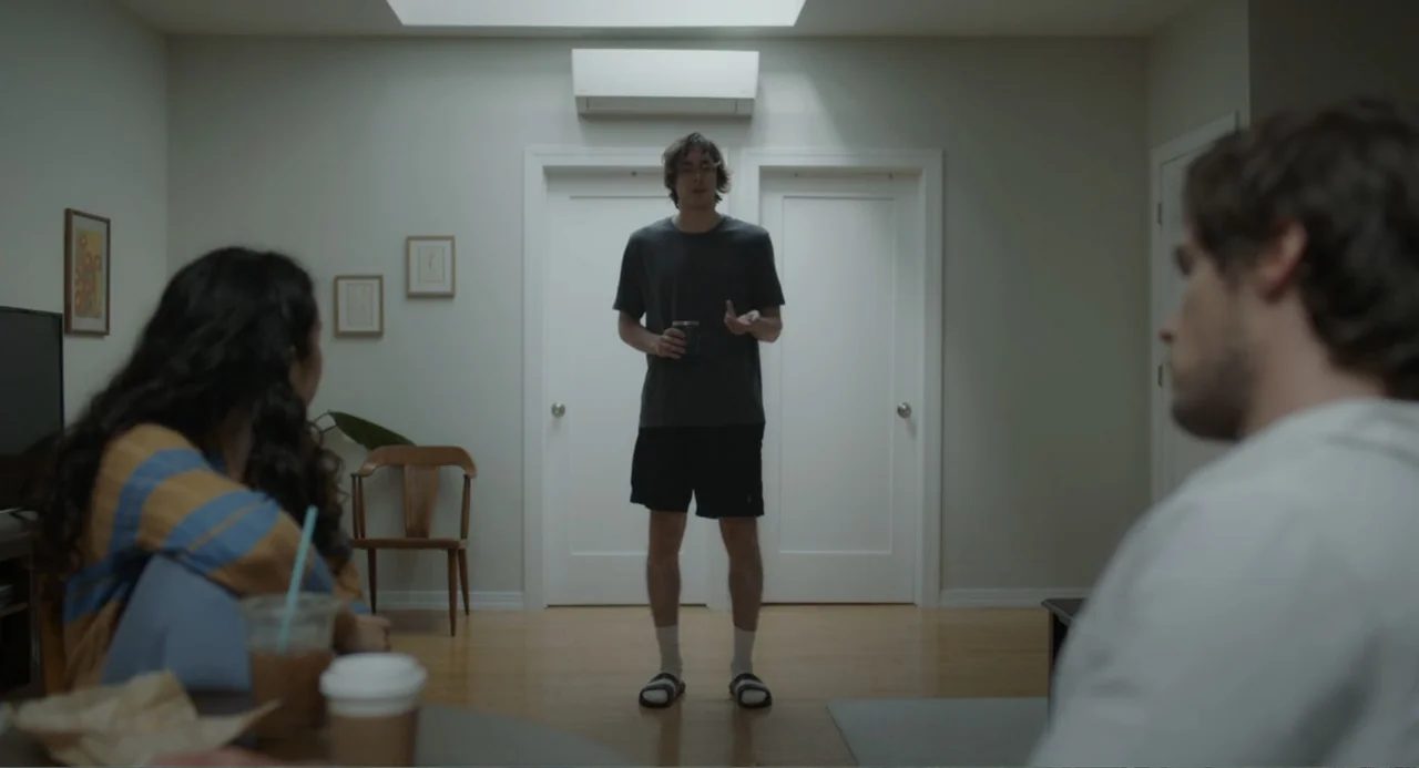 This Closeness (2024) movie still: fullbody — A modern apartment living room with white walls and a central skylight overhead. A young man stands in…