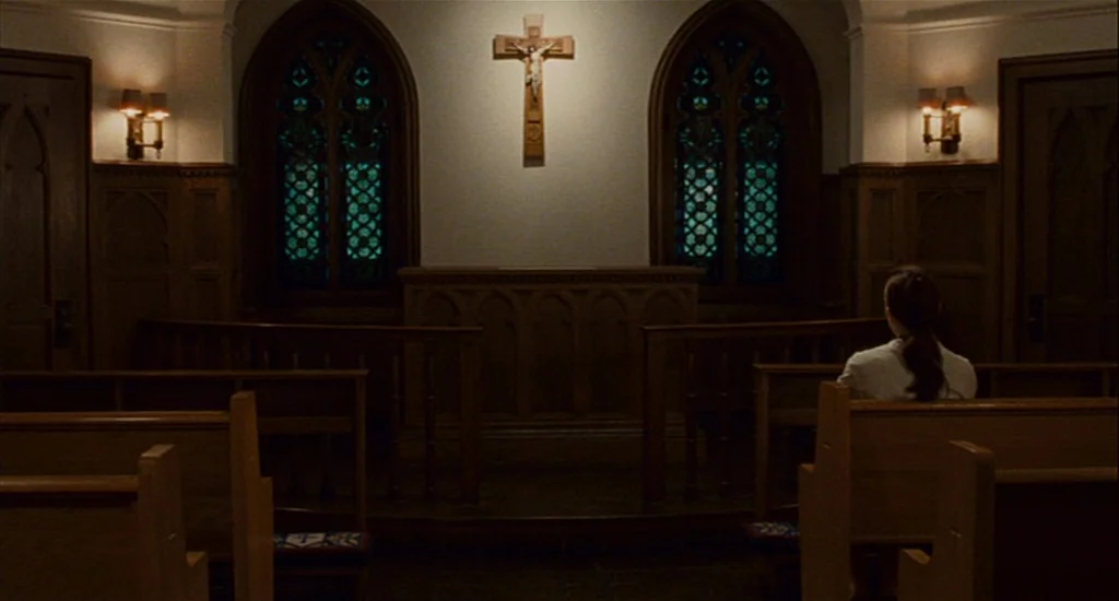 Choke (2008) movie still: wide — A dark, wood-paneled chapel features arched stained glass windows and a centered wooden crucifix. A single woman…