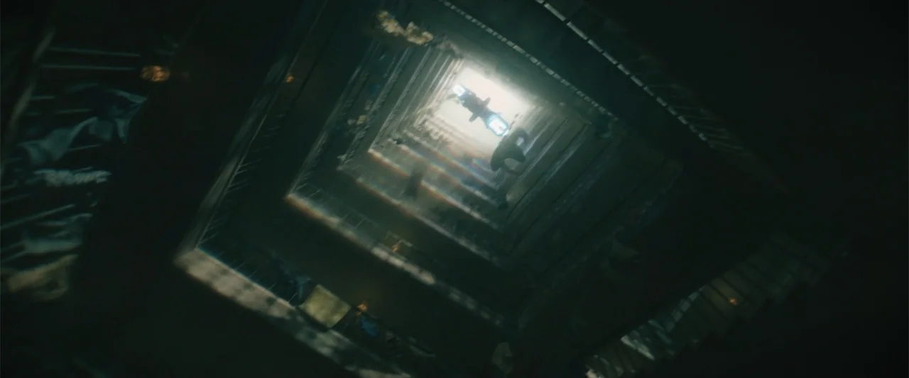 Black Adam (2022) movie still: establishing — A dark, vertical stairwell shaft descends from a single bright light source at the top. The…