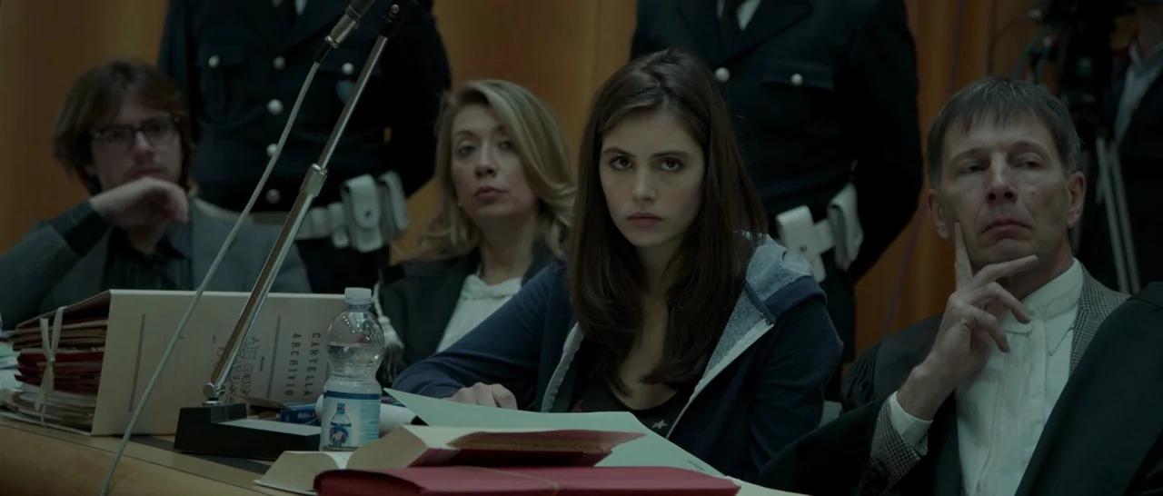 The Face of an Angel (2014) movie still: medium — A wood-paneled room serves as a formal legal setting with multiple people at desks. The central…