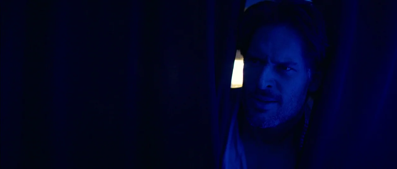 Magic Mike XXL (2015) movie still: closeup — A man peers through a gap in heavy dark curtains into a dimly lit room. The deep blue lighting masks…