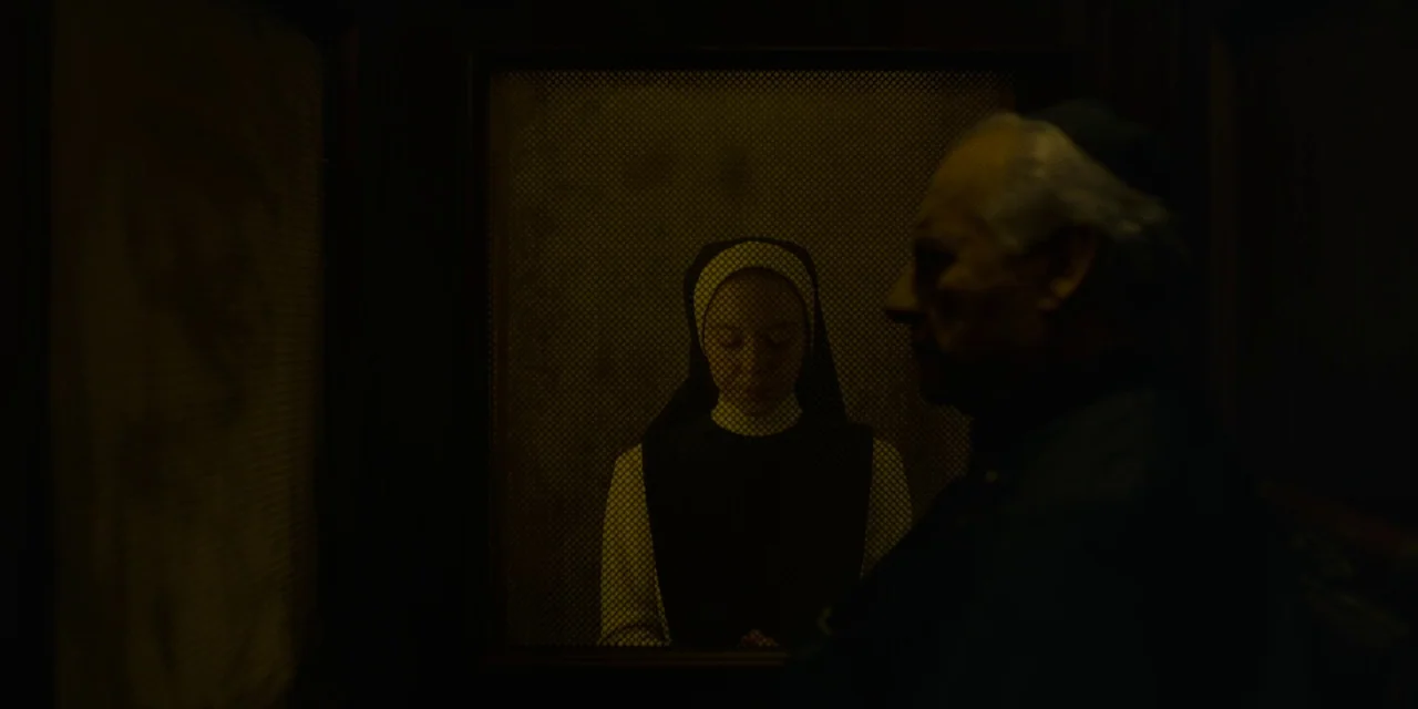 Immaculate (2024) movie still: medium — A dark, wood-paneled wooden enclosure separates two people by a fine metal mesh screen. A nun in full habit…