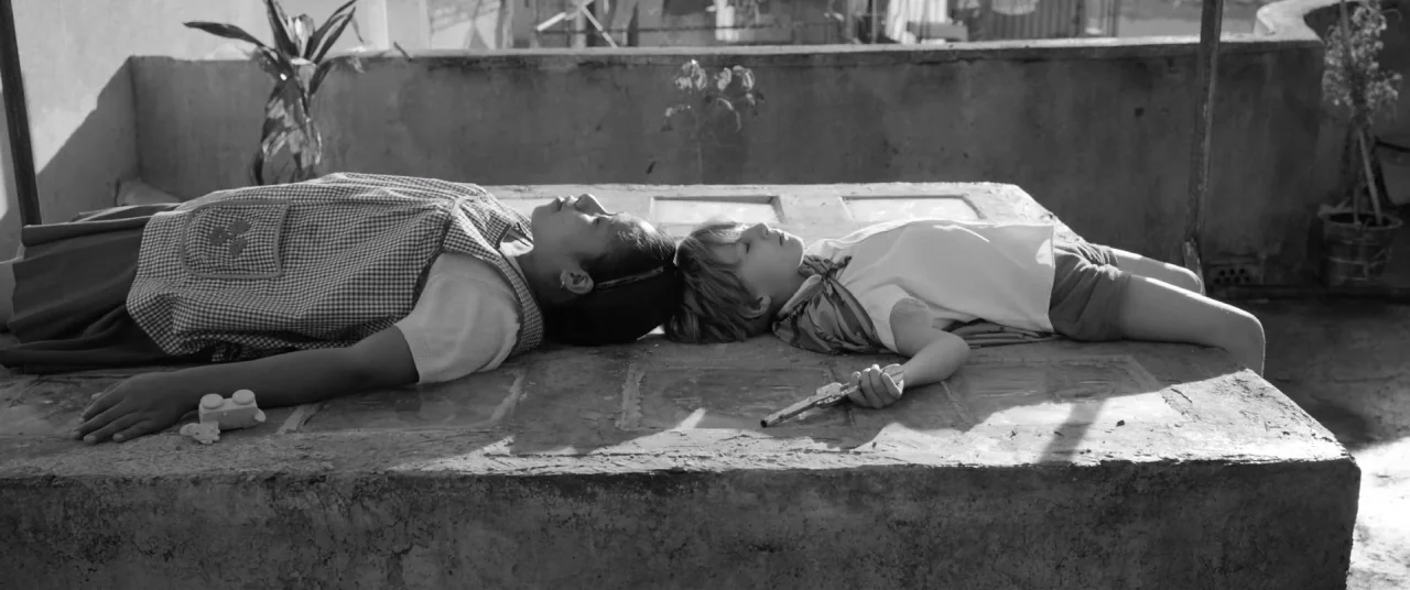 Roma (2018) movie still: medium — A flat concrete surface on a rooftop contains a young girl and boy. They lie side-by-side with heads touching…