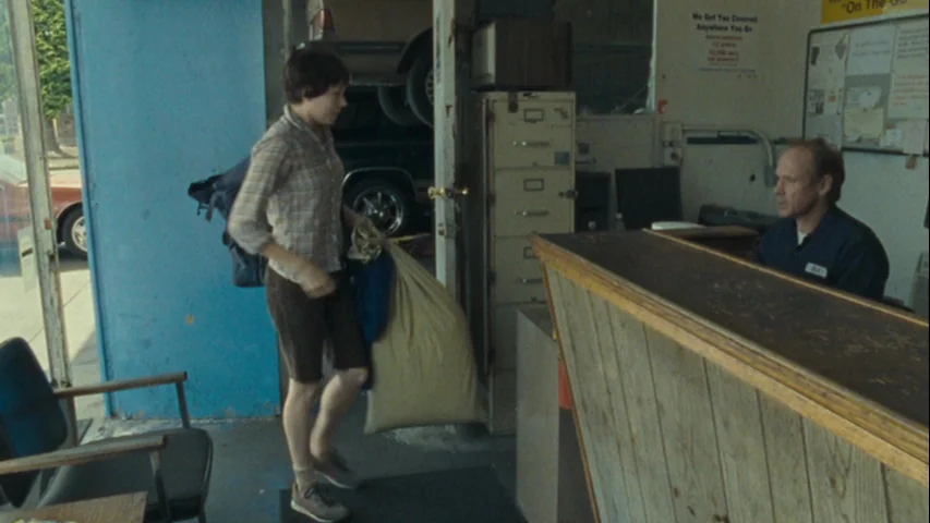 Wendy and Lucy (2008) movie still: fullbody — An auto repair shop office with a wooden counter and metal filing cabinets. A young person enters…