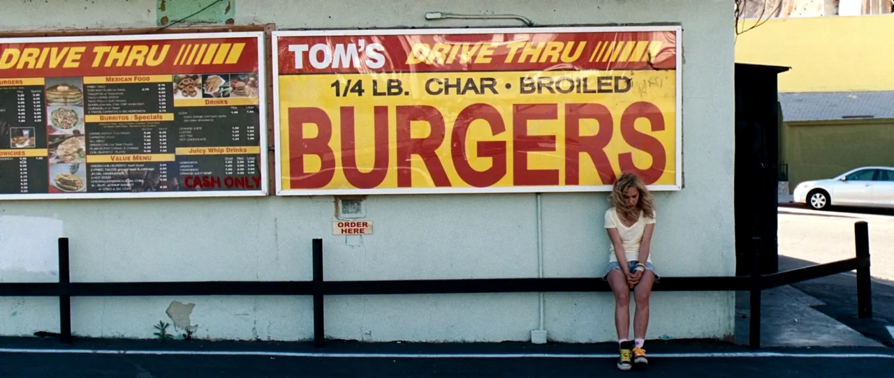 Little Birds (2011) movie still: wide — A painted stucco exterior wall features large red and yellow drive-thru menu signs. A young woman sits on a…