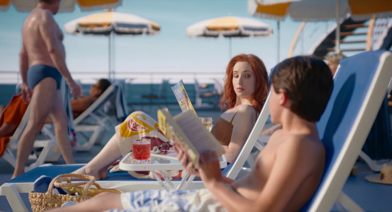 Beau Is Afraid (2023) movie still: medium — A cruise ship deck features rows of blue lounge chairs under yellow patio umbrellas. A red-haired woman…