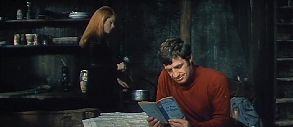 Mississippi Mermaid (1969) movie still: medium — A dimly lit room with dark wooden walls and shelves holding modest kitchenware items. A man sits at…
